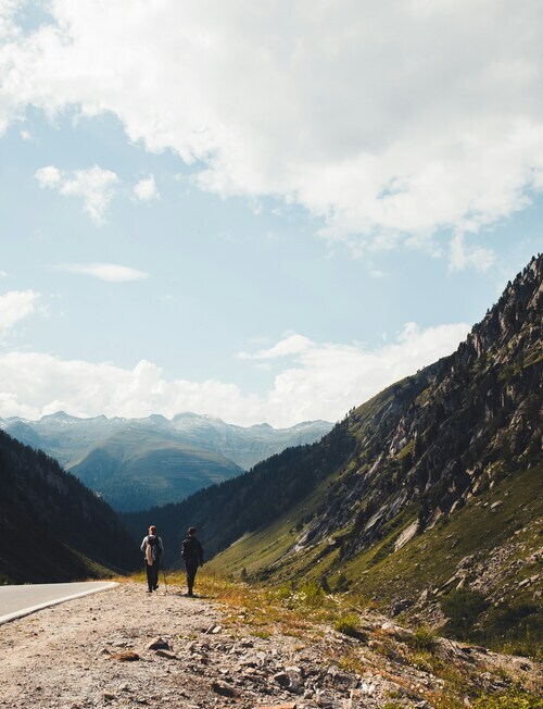 Two people walking along a mountain road overlooking a wide valley and distant peaks